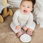 A smiling baby sitting on a neutral-toned rug, wearing a cream-colored top with striped pants, next to a white hanging ornament with a handprint impression.