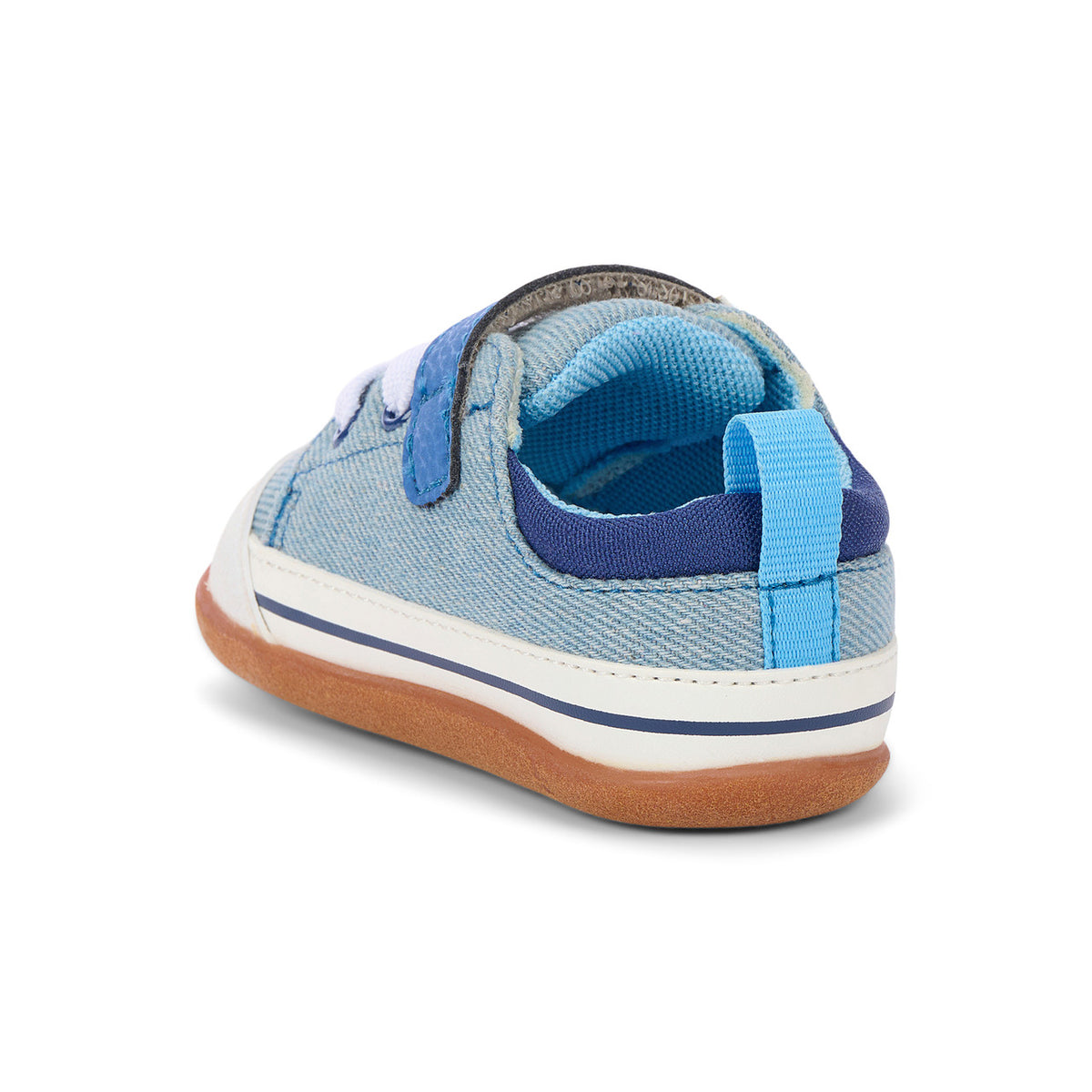 A single toddler's canvas sneaker shown from a rear three-quarter angle on a white background. The shoe features a light blue denim-like canvas upper, a dark blue elastic strap, a bright blue pull tab, a white midsole stripe, and a brown gum sole.