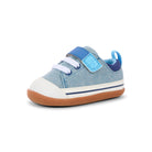 A single toddler's canvas shoe in light blue denim-like fabric, featuring a white rubber toe cap, white laces, a light blue hook-and-loop strap, a dark blue heel pull tab, and a brown gum sole, shown against a plain white background.