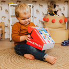 A young toddler sits on a woven rug, eagerly opening a red and white SmartMax My First People toy box.