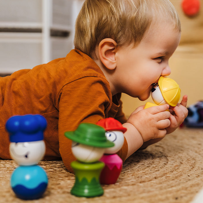 A young toddler plays on the floor with colorful SmartMax My First People magnetic figures, holding a yellow construction hat toy piece.
