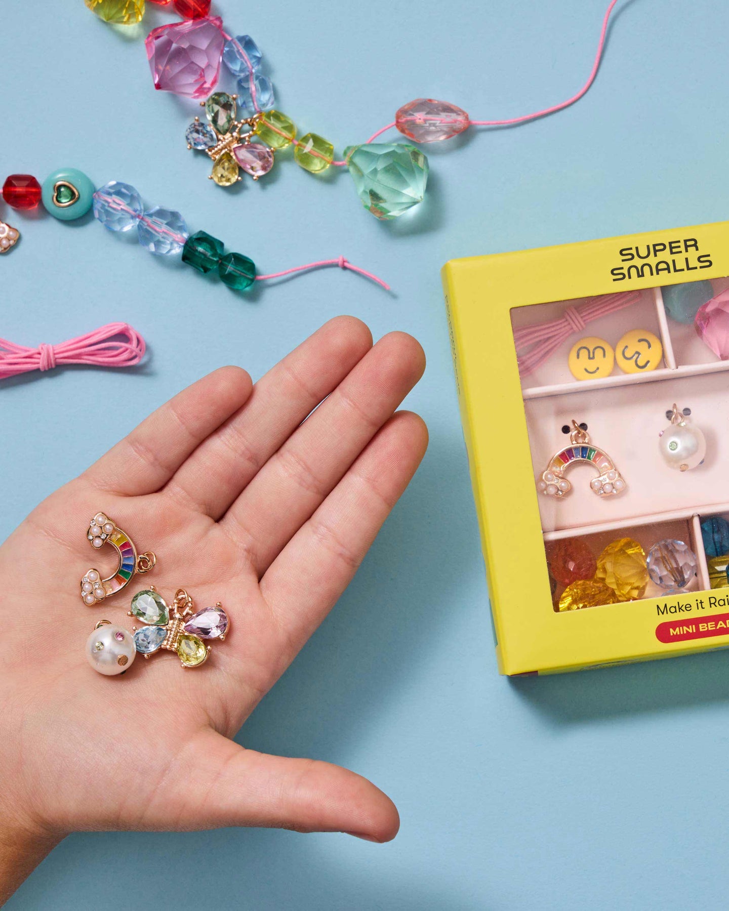 Colorful jewelry set with a hand holding a small piece, next to a 'Super Smalls' packaging on a blue background.