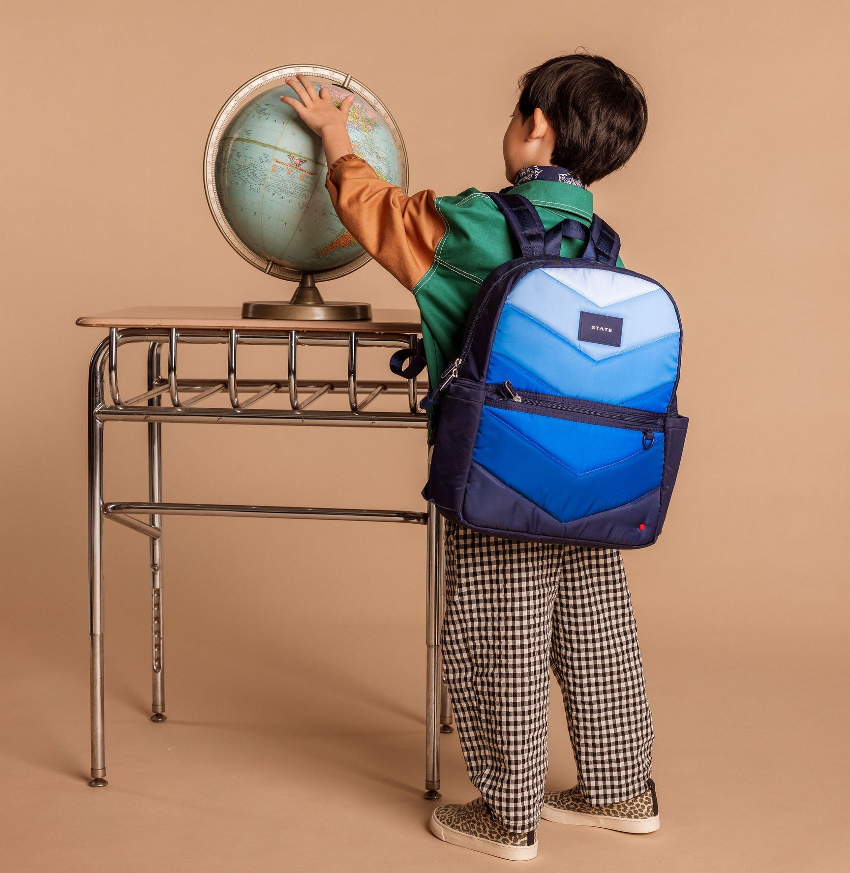 Child with a blue backpack looking at a globe on a desk against a beige background
