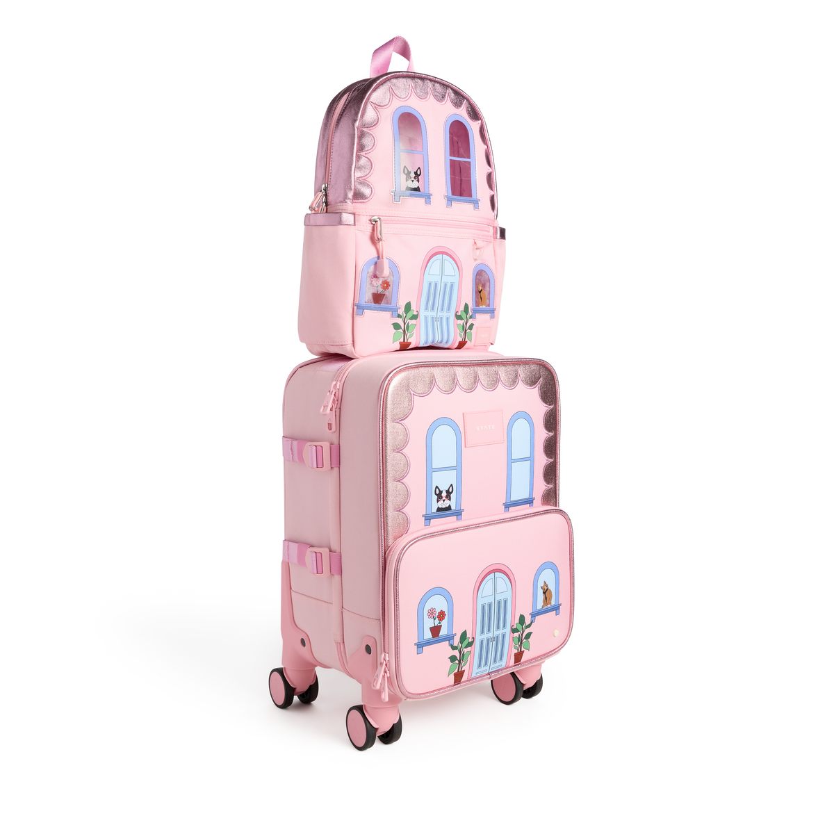 A pink State Bags Mini Logan house-style backpack stacked on top of a matching pink rolling carry-on suitcase, featuring blue telescopic handles, 360-degree spinner wheels, and front graphic details including arched windows, potted plants, and a central blue front door against a transparent background.