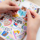 Pets Stickers by Eeboo in use, with hands peeling a cute hamster sticker reading a book from a colorful sticker sheet featuring various illustrated animal characters, including a mouse with cookies, a snail, and a cat.