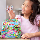 A smiling young girl holding up cut-out pet stickers while seated next to the colorful arched packaging of Pets Stickers by Eeboo.