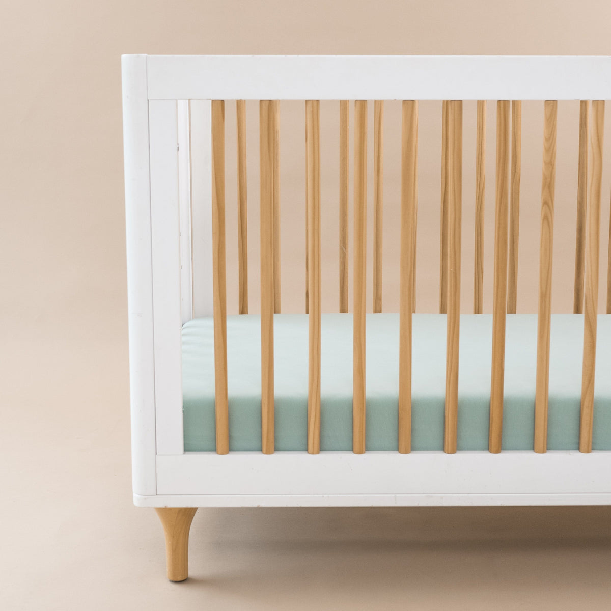 White crib with wooden slats and a light green mattress against a beige background