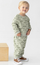 A toddler standing in a studio wearing a sage green long-sleeve sweatshirt and matching sweatpants with a repeating fish print, paired with brown sneakers. A light wood block is visible on the left side of the frame.