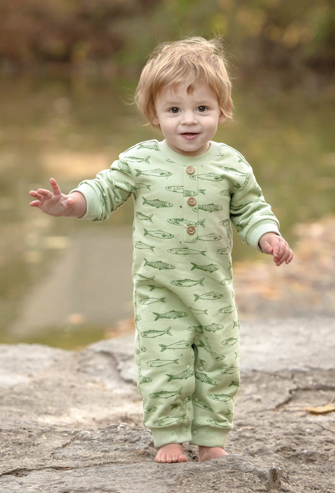 A toddler standing barefoot on a rock outdoors, wearing a light sage green long-sleeve footed sleeper with an allover fish print and brown buttons on the chest.