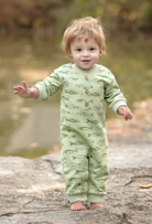 A toddler standing barefoot on a rock outdoors, wearing a light sage green long-sleeve footed sleeper with an allover fish print and brown buttons on the chest.