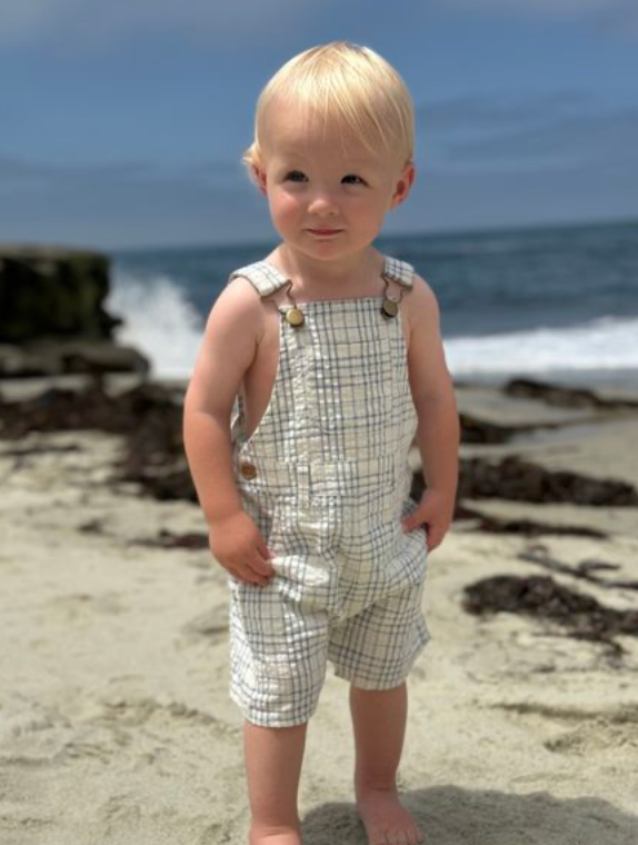 Child in checkered overalls standing on a beach with ocean in the background