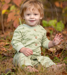 A smiling toddler sitting outdoors in grass and fallen leaves, wearing a light sage green one-piece outfit with fish print and three brown buttons.