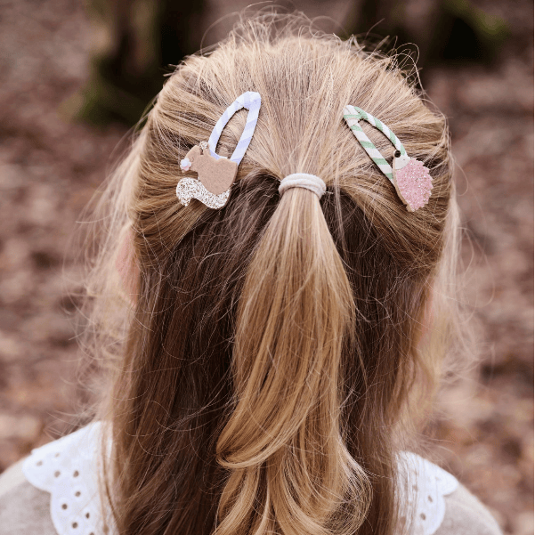 A close-up back view of a child with blonde hair in a low ponytail, wearing two decorative hair clips in an outdoor setting with a blurred brown background.