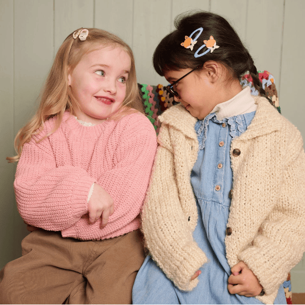 Two young girls sitting side-by-side wearing cozy knit clothing. The girl on the left wears a pink cable-knit sweater and brown pants, while the girl on the right wears a cream chunky-knit cardigan over a light blue denim dress.