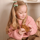 A young girl with long blonde hair wearing a pink knit sweater and a green hair clip, holding a piece of brown bread while sitting in a cozy indoor setting.