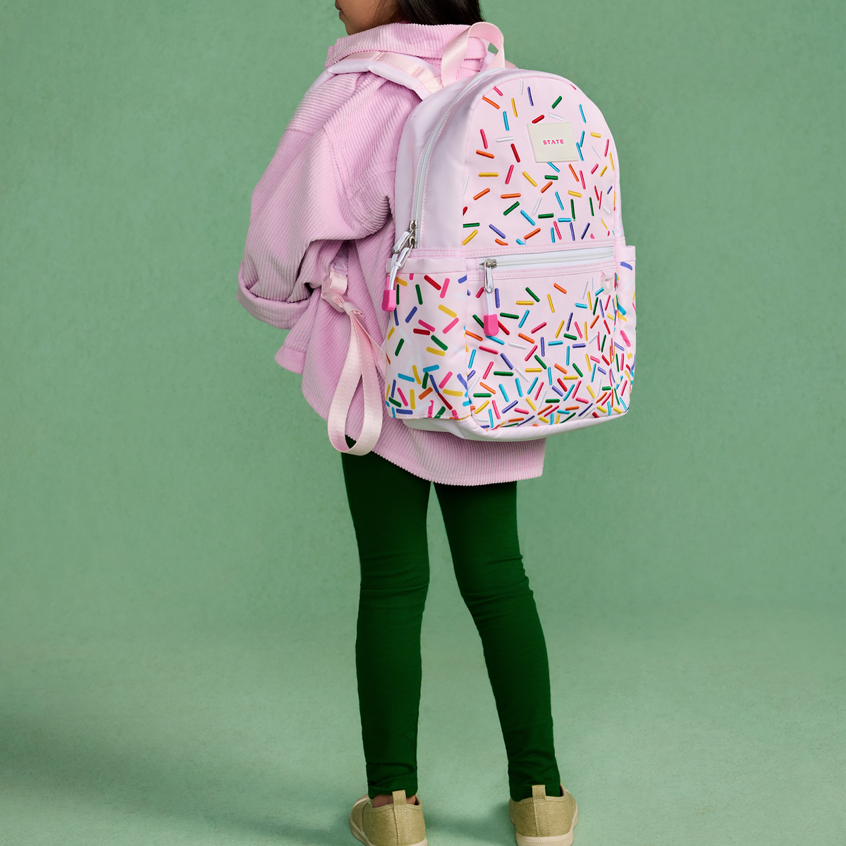 Person wearing a pink backpack with colorful sprinkles pattern on a green background