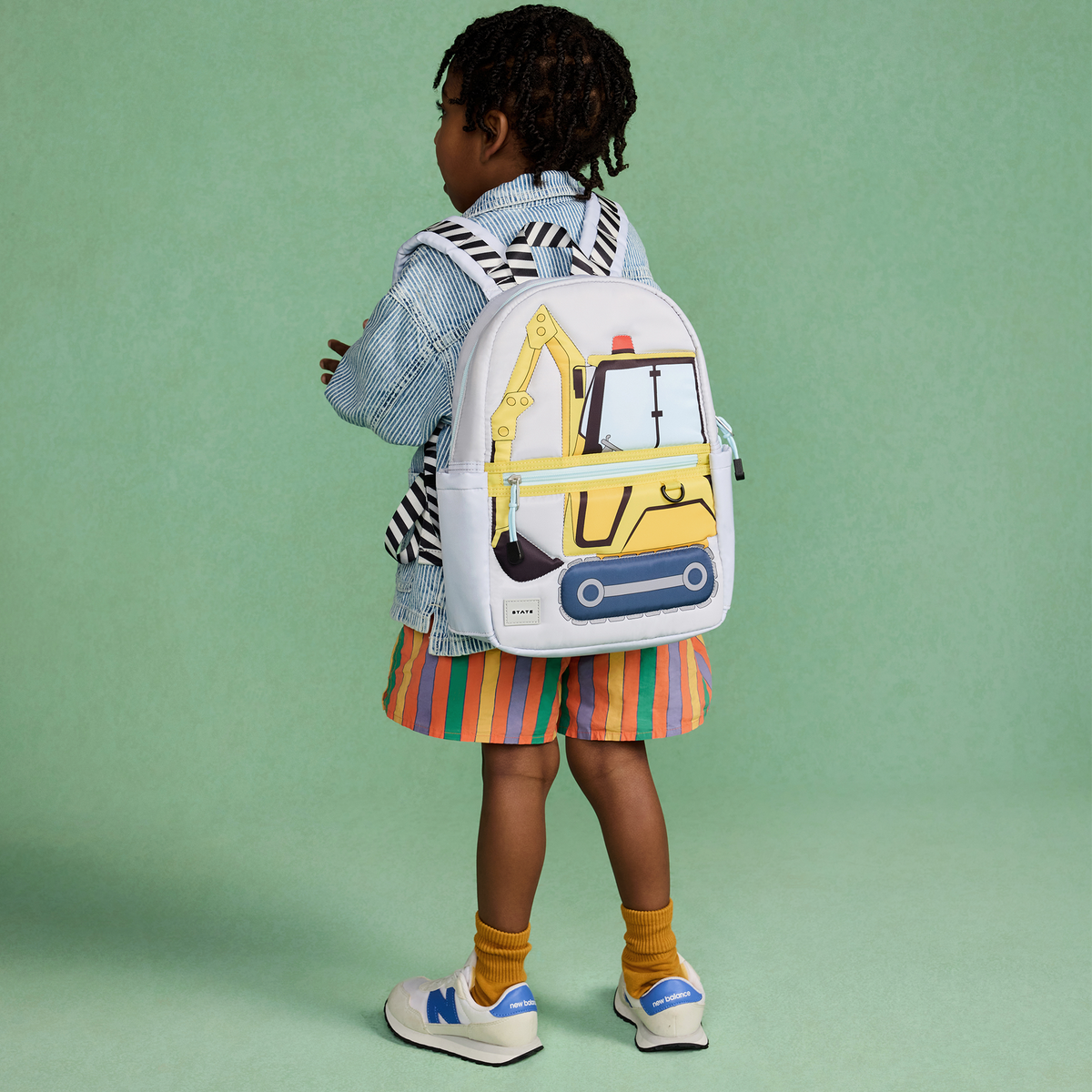 A child wearing a white backpack with a yellow construction excavator design, standing against a solid mint green background.
