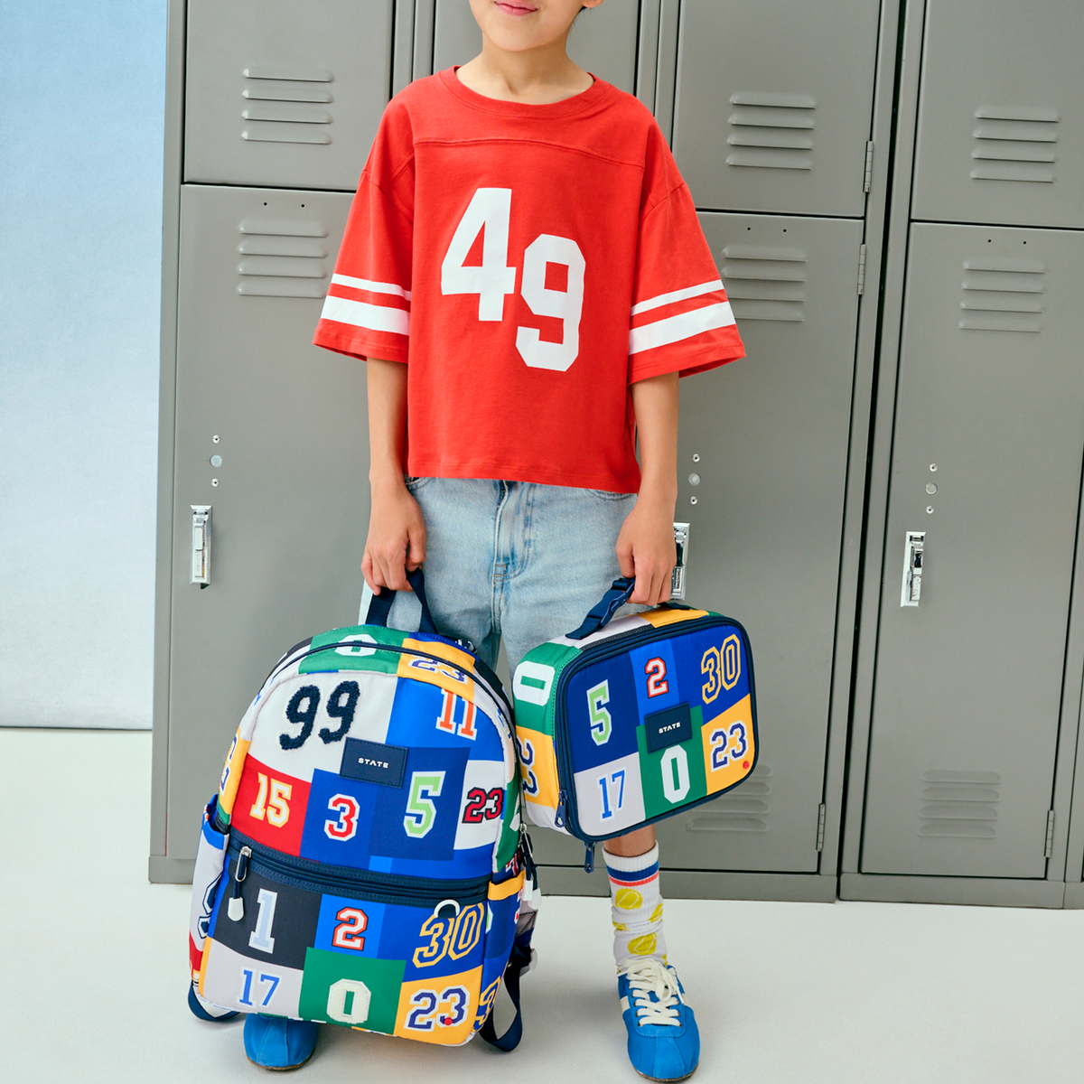 Child holding two colorful backpacks with numbers in front of gray lockers.