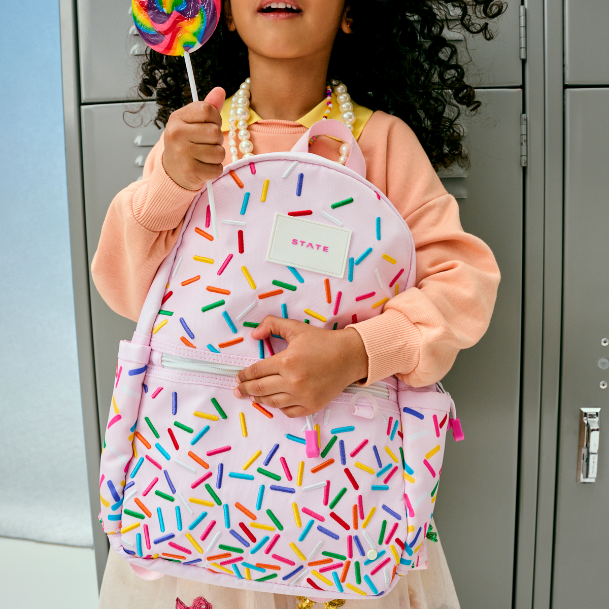 Child holding a backpack with colorful sprinkles pattern and a lollipop in front of lockers.