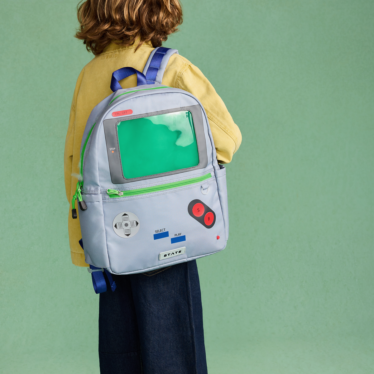 A child wearing a yellow shirt and dark pants is shown from behind, carrying a light blue backpack designed to look like a retro video game console with a green screen, green zipper trim, and red buttons. The background is a solid sage green studio backdrop.