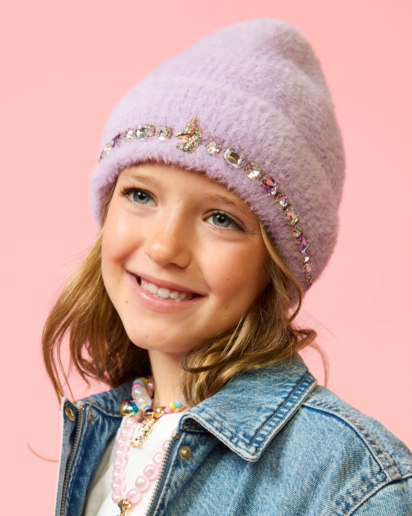 A young girl smiling while wearing a fuzzy lavender knit beanie with a decorative band of rhinestones and a butterfly embellishment, paired with a denim jacket against a solid pink background.