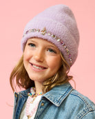 A young girl smiling while wearing a fuzzy lavender knit beanie with a decorative band of rhinestones and a butterfly embellishment, paired with a denim jacket against a solid pink background.
