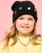 Child wearing a black beanie with colorful embellishments on a pink background
