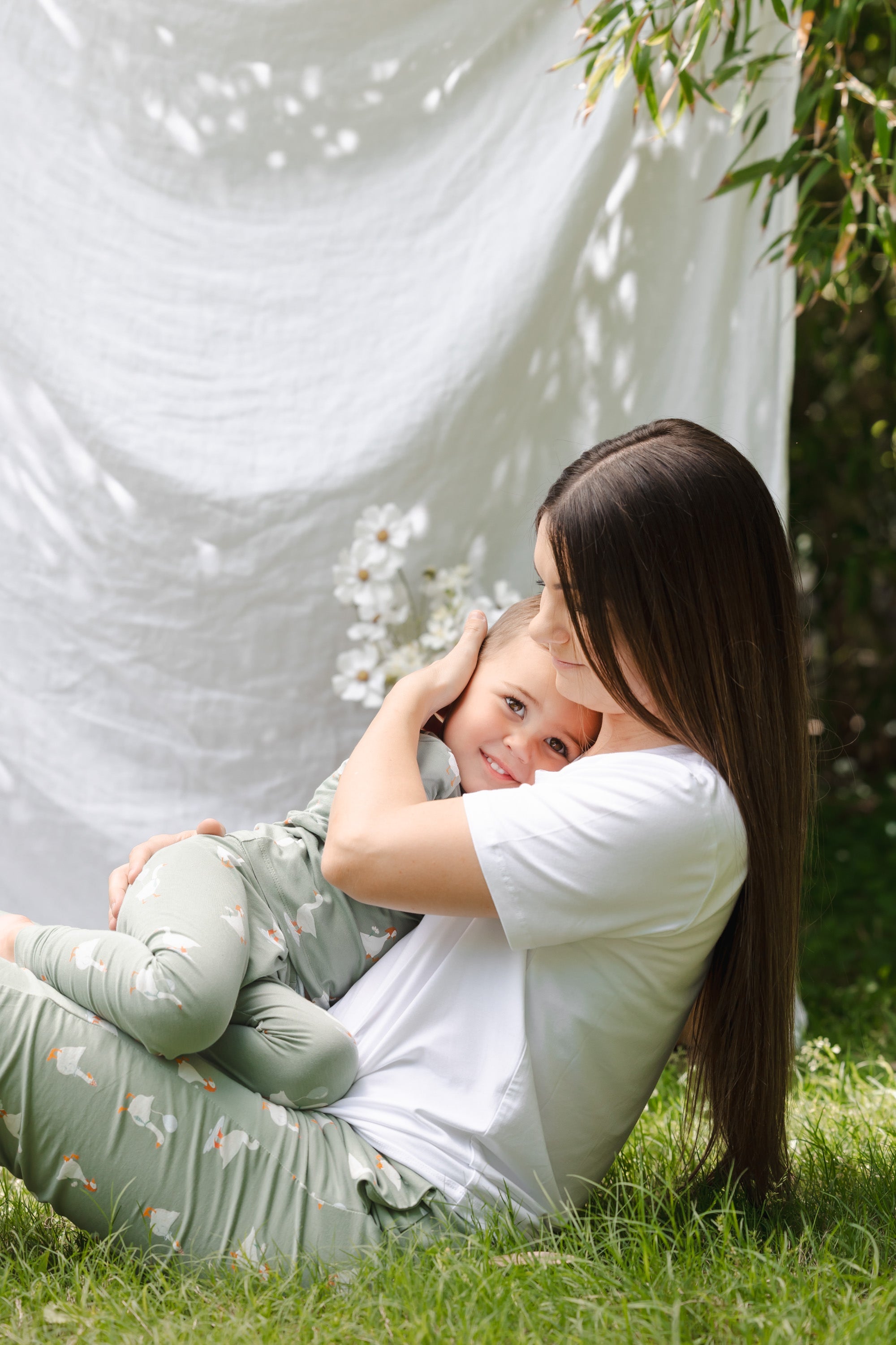 Woman sitting on grass with a child, both wearing light-colored clothing, with a white curtain and greenery in the background.