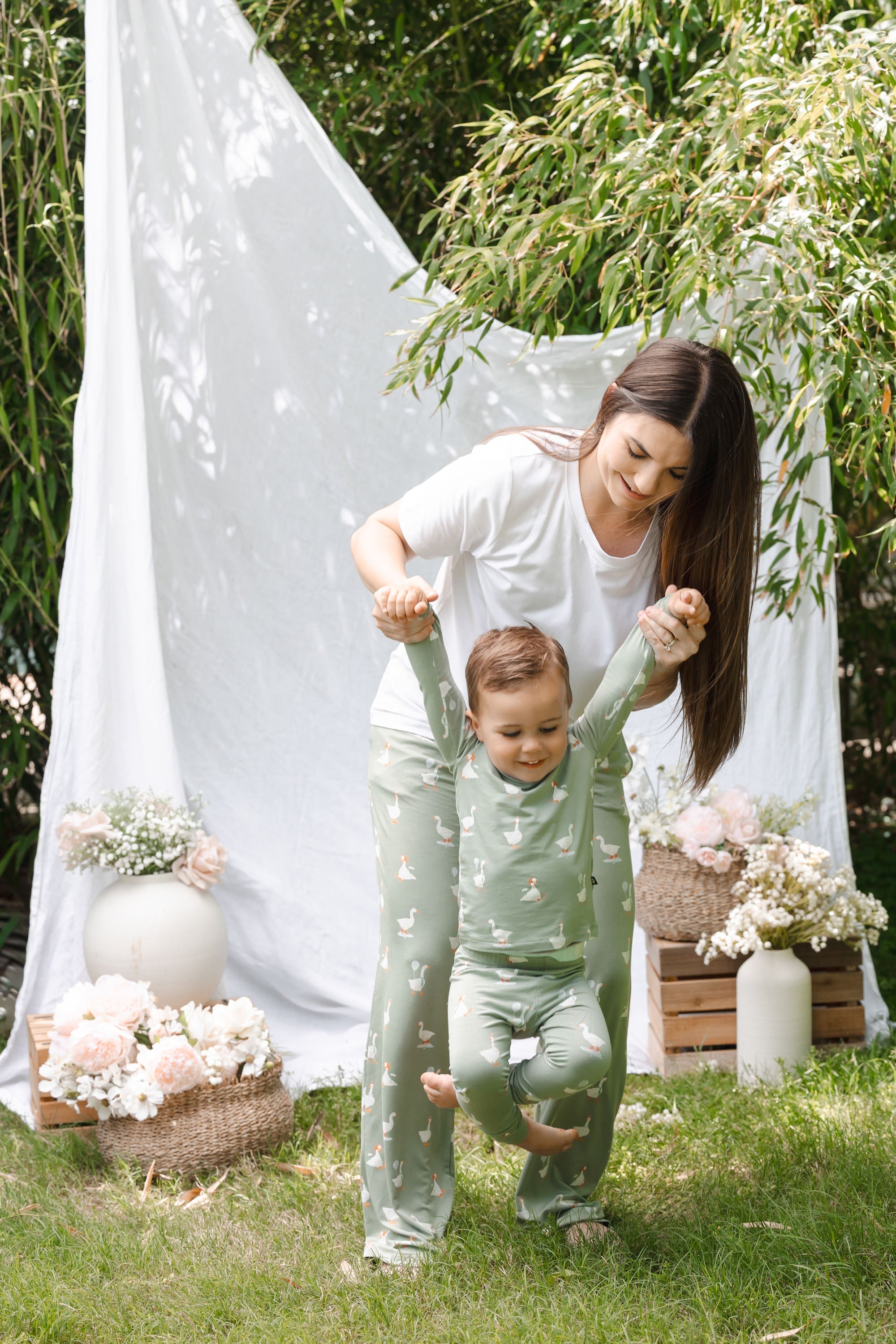 Woman and child in matching outfits standing outdoors with flowers and a white curtain in the background