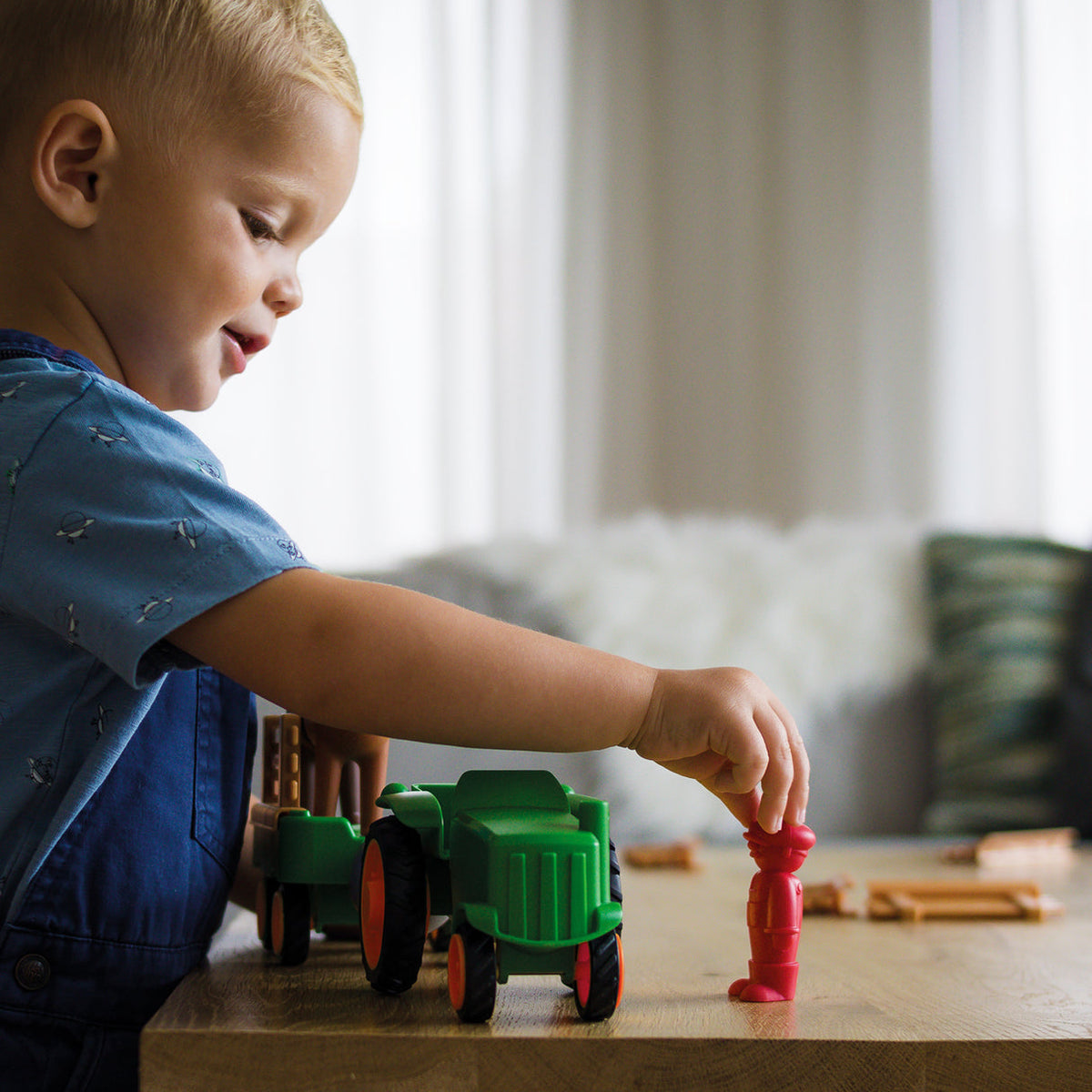 A young blond boy playing on the floor, smiling as he places a red toy figure near the green Smartmax My First Farm Tractor set.
