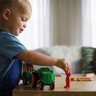 A young blond boy playing on the floor, smiling as he places a red toy figure near the green Smartmax My First Farm Tractor set.