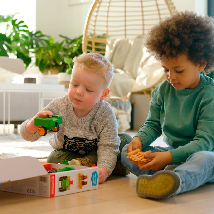Two young children playing on the floor together with the green Smartmax My First Farm Tractor set, assembling the magnetic pieces near the open product box.
