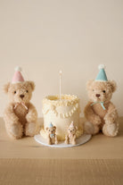 Three teddy bears with birthday hats around a small cake with a candle on a wooden surface.