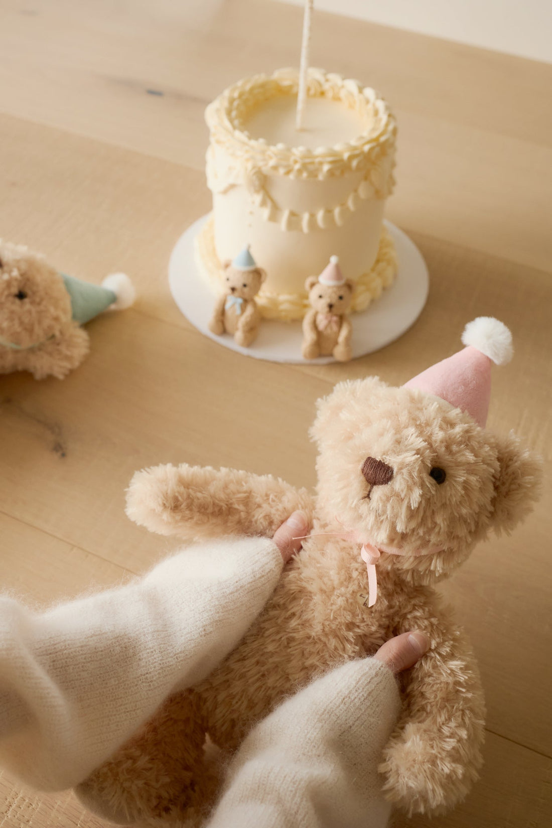 Teddy bear with a pink party hat being held, with a small cake and teddy bears in the background on a wooden surface.