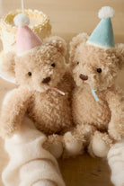 Two teddy bears wearing party hats sitting on a white blanket.