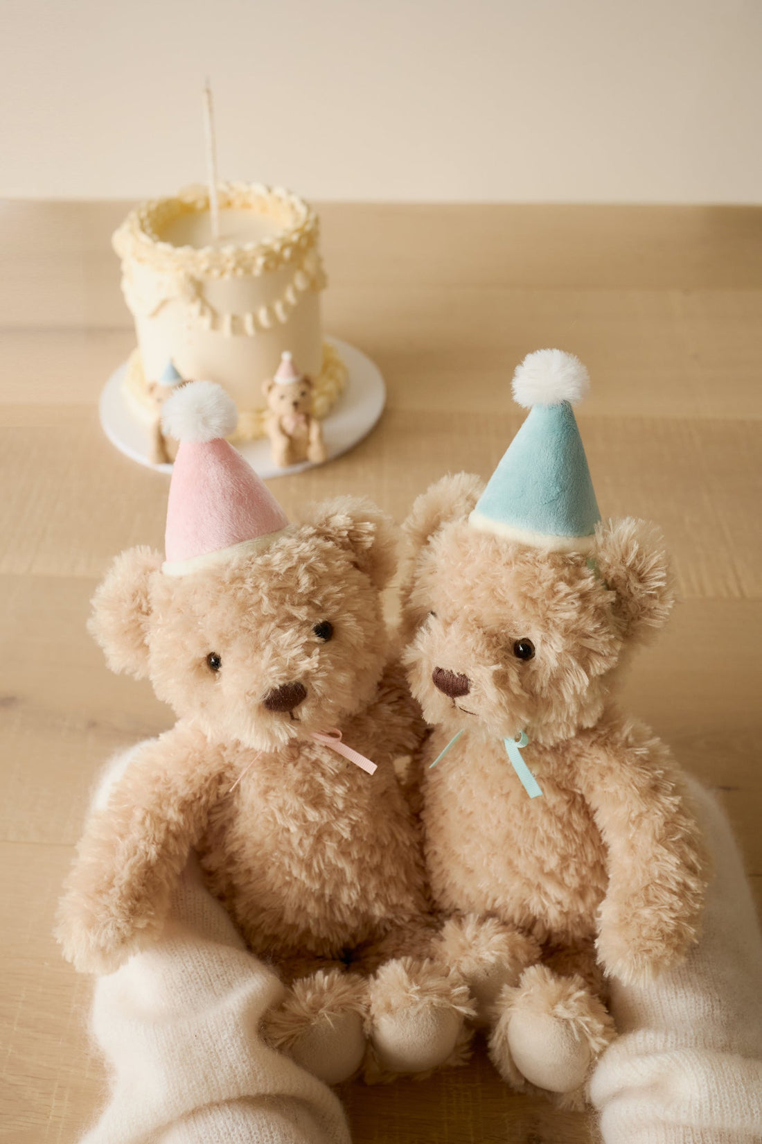 Two teddy bears wearing party hats sitting on a wooden surface with a small cake in the background.