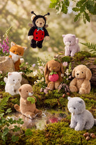 A collection of Jamie Kay plush toys—including a ladybug, a fox, a teddy bear, a bunny, a puppy, and a grey kitty—arranged together in a natural outdoor woodland setting near a stream.