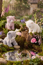 A pink plush animal, a grey kitten, and a white plush animal posed together in an enchanted woodland setting with moss, rocks, and blooming flowers near a reflective pool.