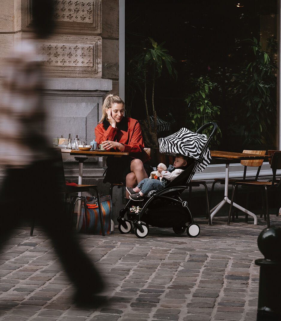 Woman sitting at an outdoor cafe table with a child in a stroller, blurred pedestrian in foreground