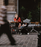 Woman sitting at an outdoor cafe table with a child in a stroller, blurred pedestrian in foreground