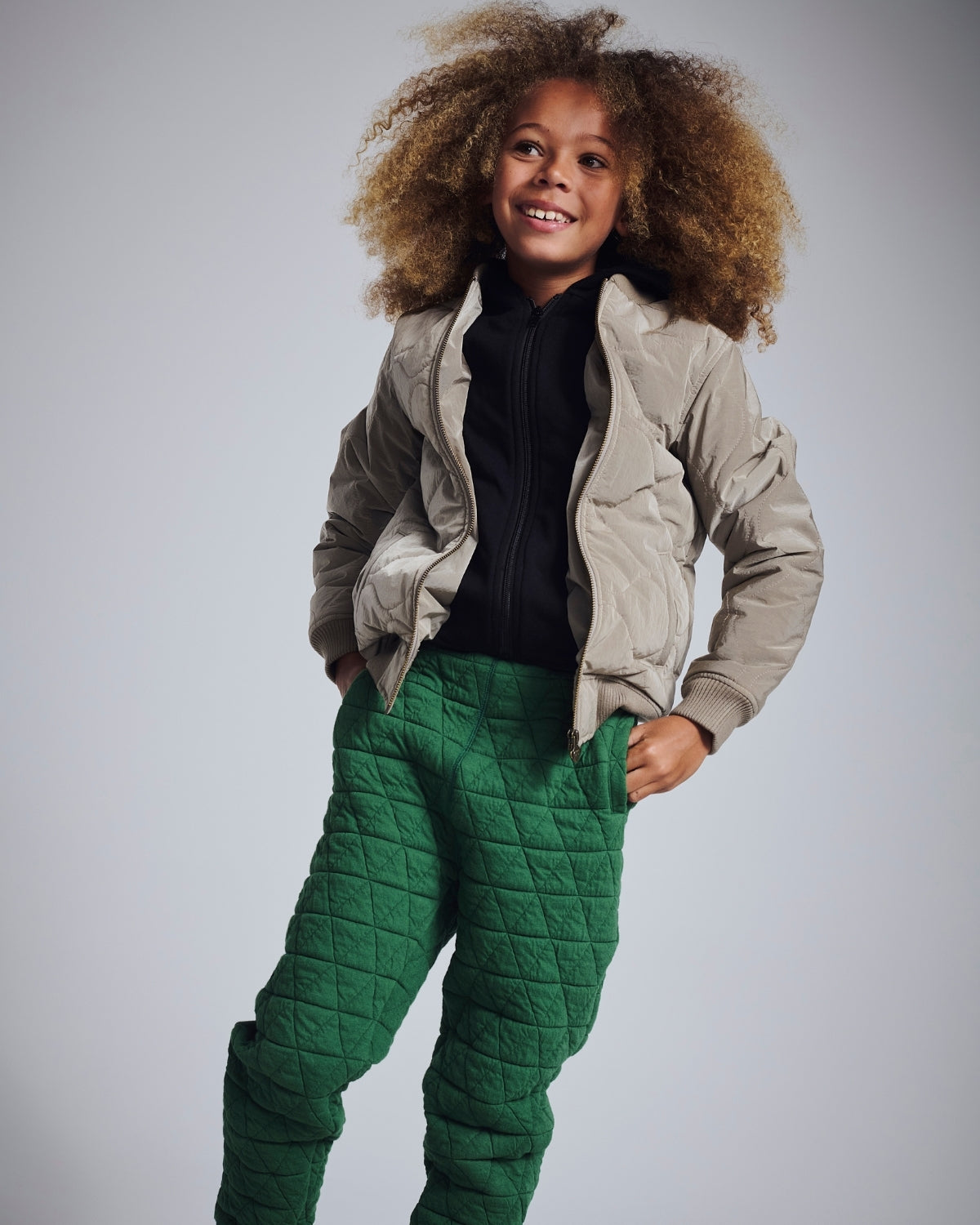 Child wearing a beige puffer jacket and green quilted pants on a gray background