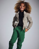 Child wearing a beige puffer jacket and green quilted pants on a gray background