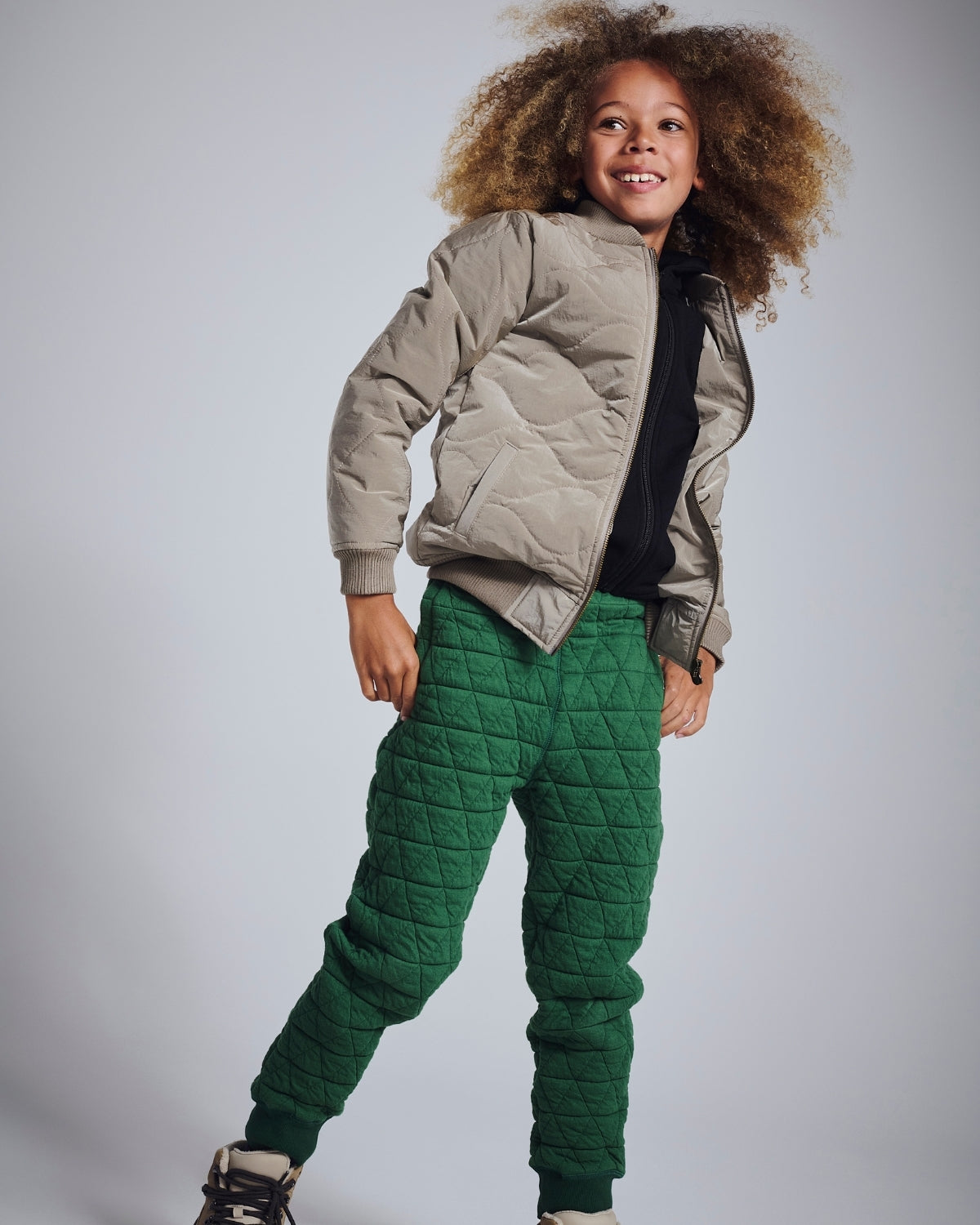 Child wearing a beige quilted jacket and green pants on a plain background