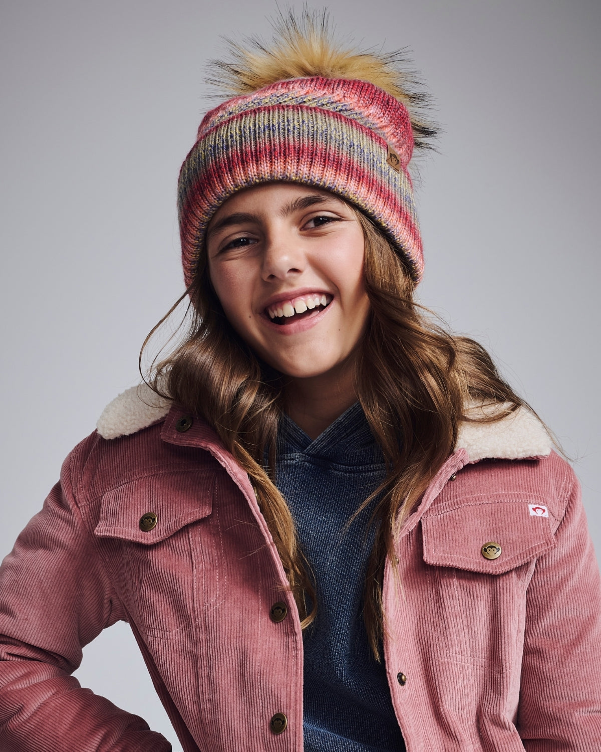 Young girl wearing a pink coat and colorful knit hat with pom-pom on a gray background