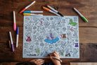 A child's coloring page with black line art illustrations and numbers, being drawn on by a hand using a marker, placed on a rustic wooden table with several colored markers around it.