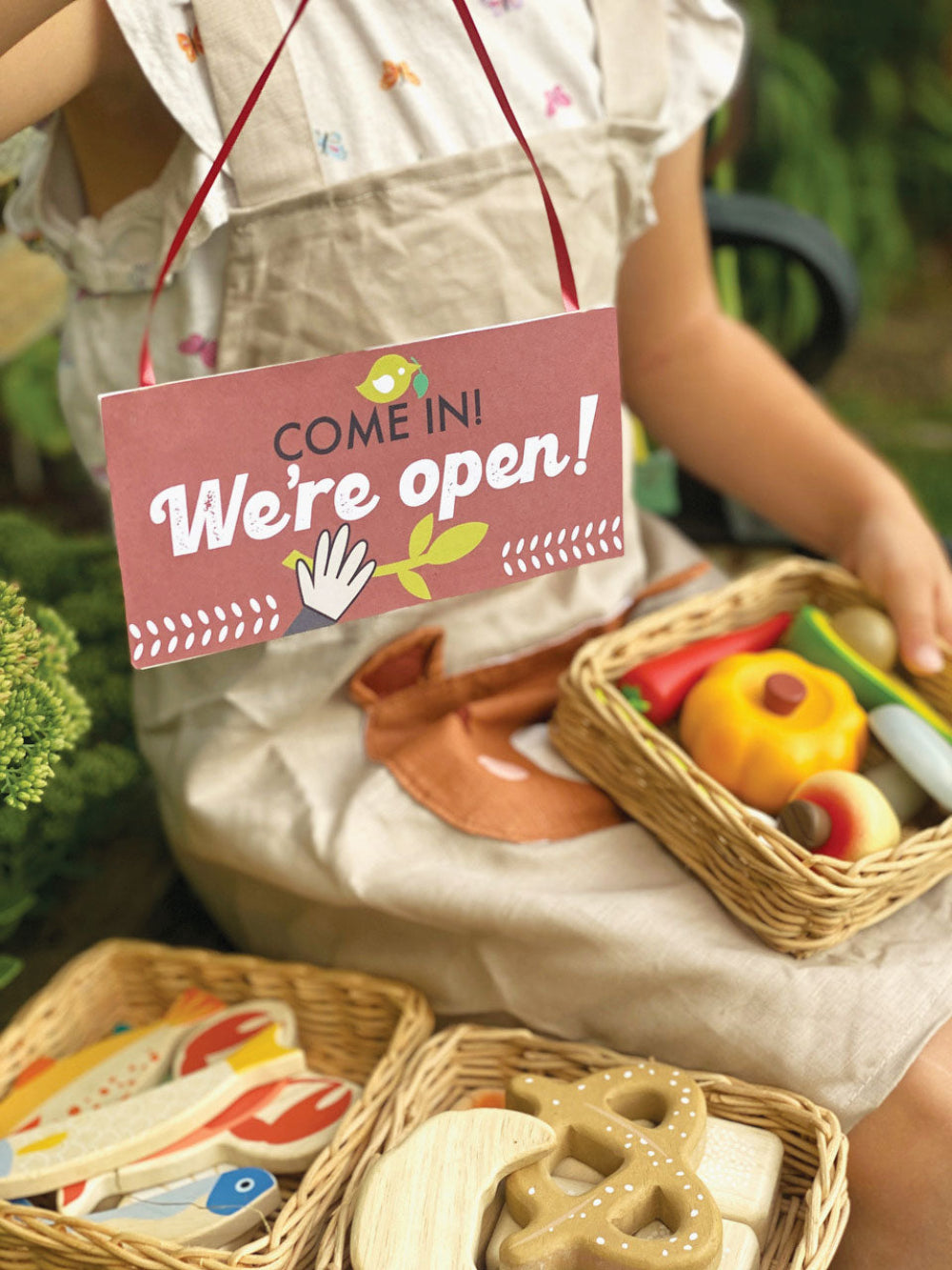 Person wearing an apron with a 'Come in! We're open!' sign and toy food items in baskets.