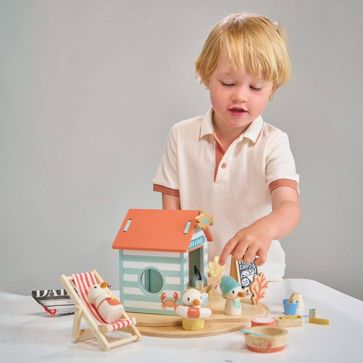 Child playing with a wooden toy house and accessories on a white surface.