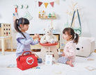 Two children playing with a red toy first aid kit in a room with toys and furniture.