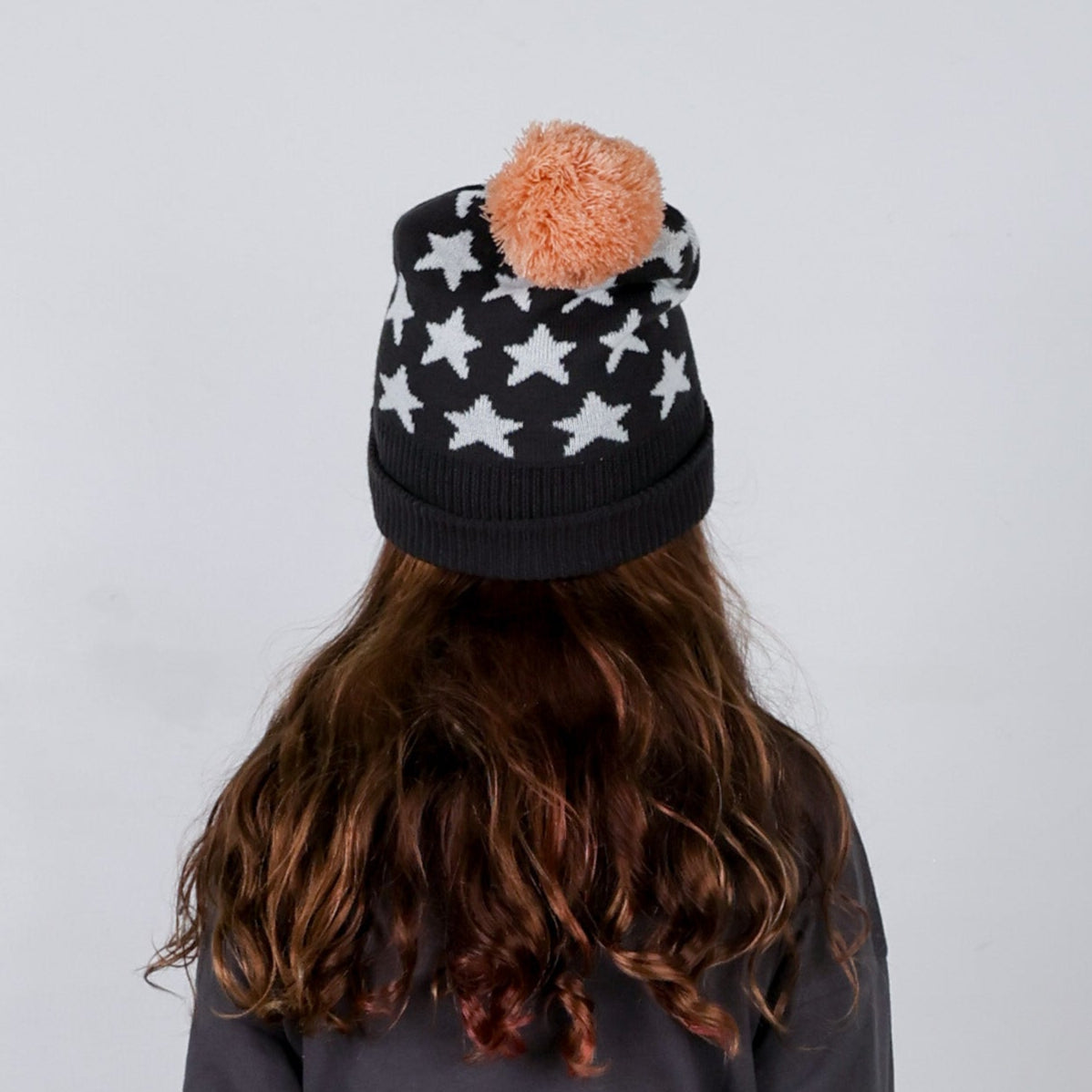 Person wearing a black beanie with white star patterns and a pink pom-pom on a plain background