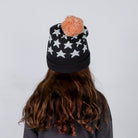 Person wearing a black beanie with white star patterns and a pink pom-pom on a plain background